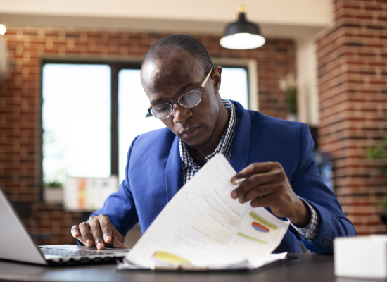 Closeup of black man seated at office desk, deeply focused on analyzing financial documents in brick wall room. African american male manager examining graphs on clipboard with laptop nearby.